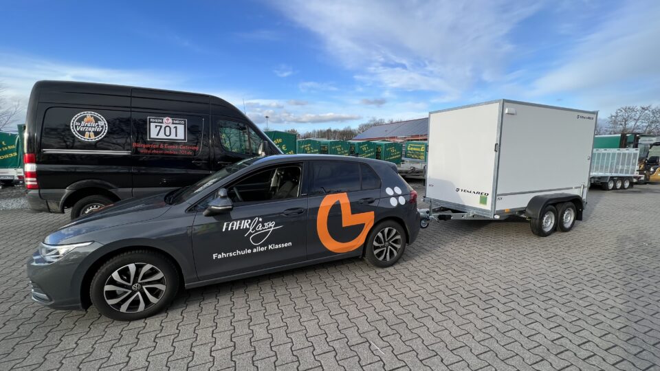 Dunkles Fahrschulauto der Fahrschule FAHRlässig mit großem Anhänger auf einem Parkplatz, beklebt mit Logo und Schriftzug Fahrschule aller Klassen