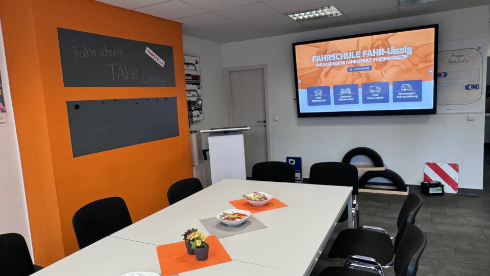 Moderner Unterrichtsraum der Fahrschule mit großem Bildschirm, Tischen, Stühlen und orangefarbener Wandgestaltung
