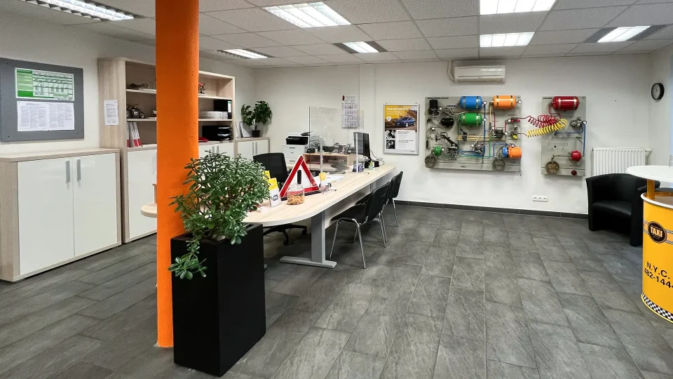 Ein Büro- und Empfangsbereich einer Fahrschule mit modernen Möbeln, Informationsmaterial, einer technischen Lehrwand für Bremssysteme und dekorativen Elementen wie Pflanzen.