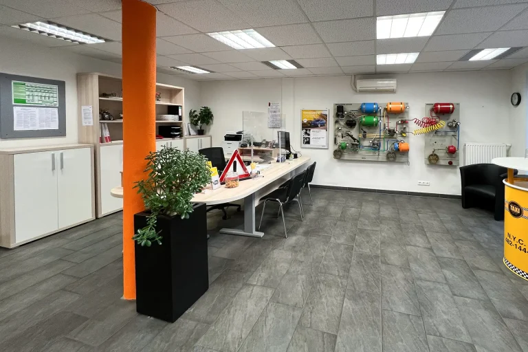 Ein Büro- und Empfangsbereich einer Fahrschule mit modernen Möbeln, Informationsmaterial, einer technischen Lehrwand für Bremssysteme und dekorativen Elementen wie Pflanzen.