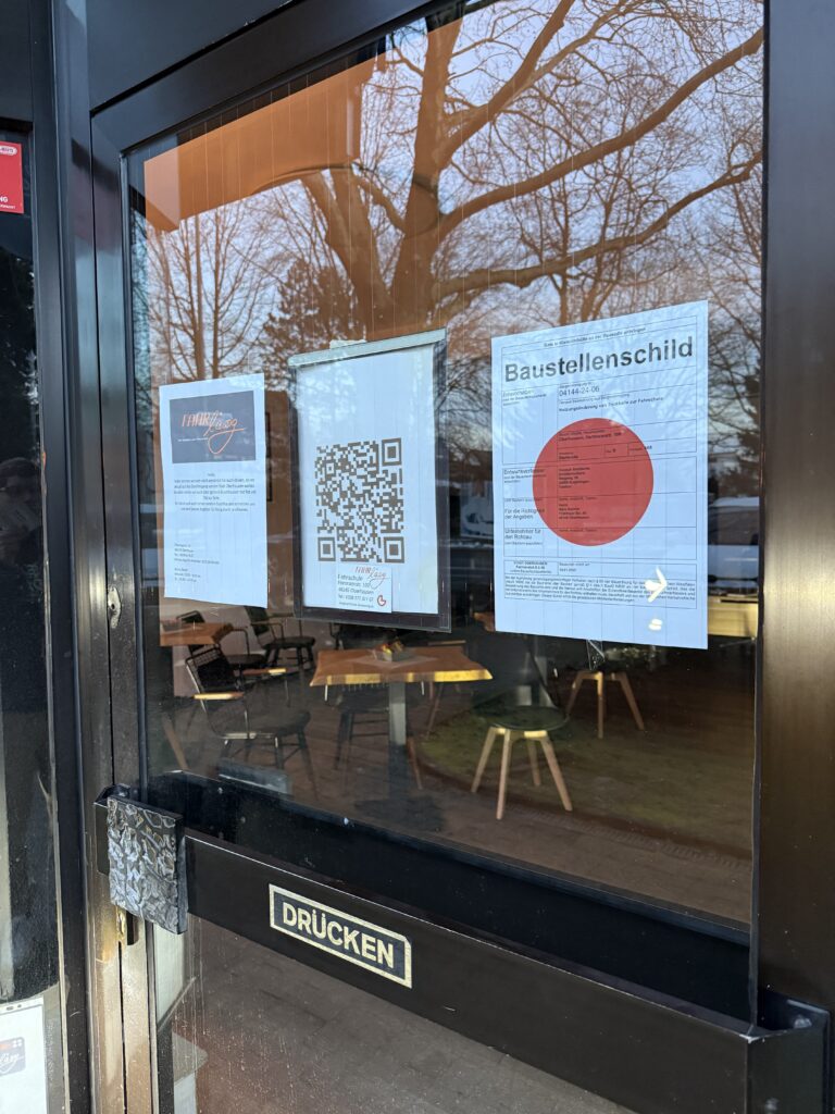 Eingangstür der Fahrschule mit Aushängen zur Neueröffnung, darunter ein QR-Code, ein Baustellenschild und Infos zur Fahrschule