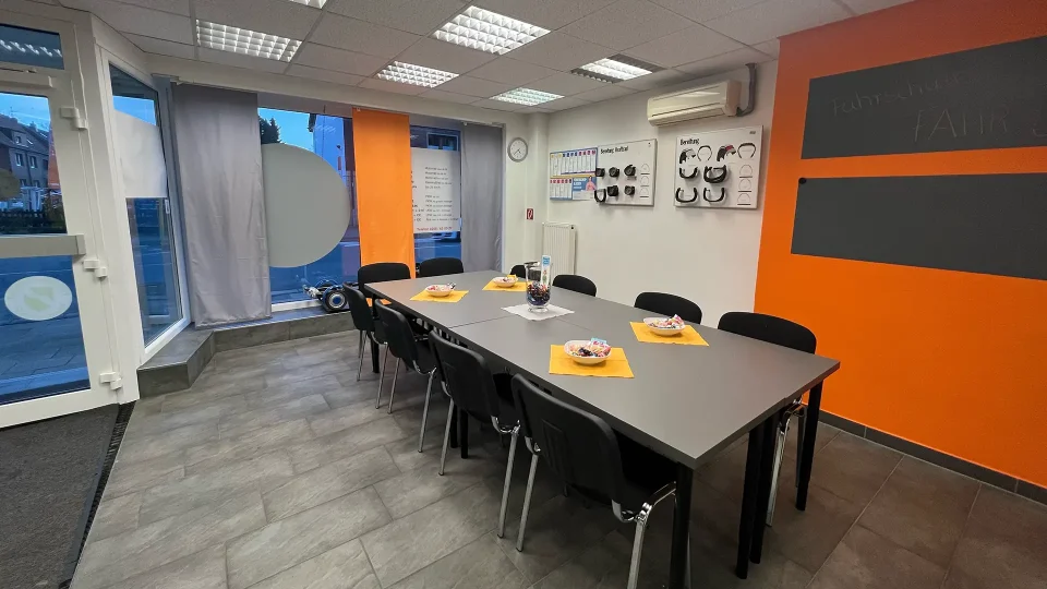 Ein moderner Unterrichtsraum einer Fahrschule mit einem zentralen Konferenztisch, mehreren Stühlen, einem Lehrerpult, Whiteboard und Flipchart, umgeben von Lernmaterialien und Informationspostern.
